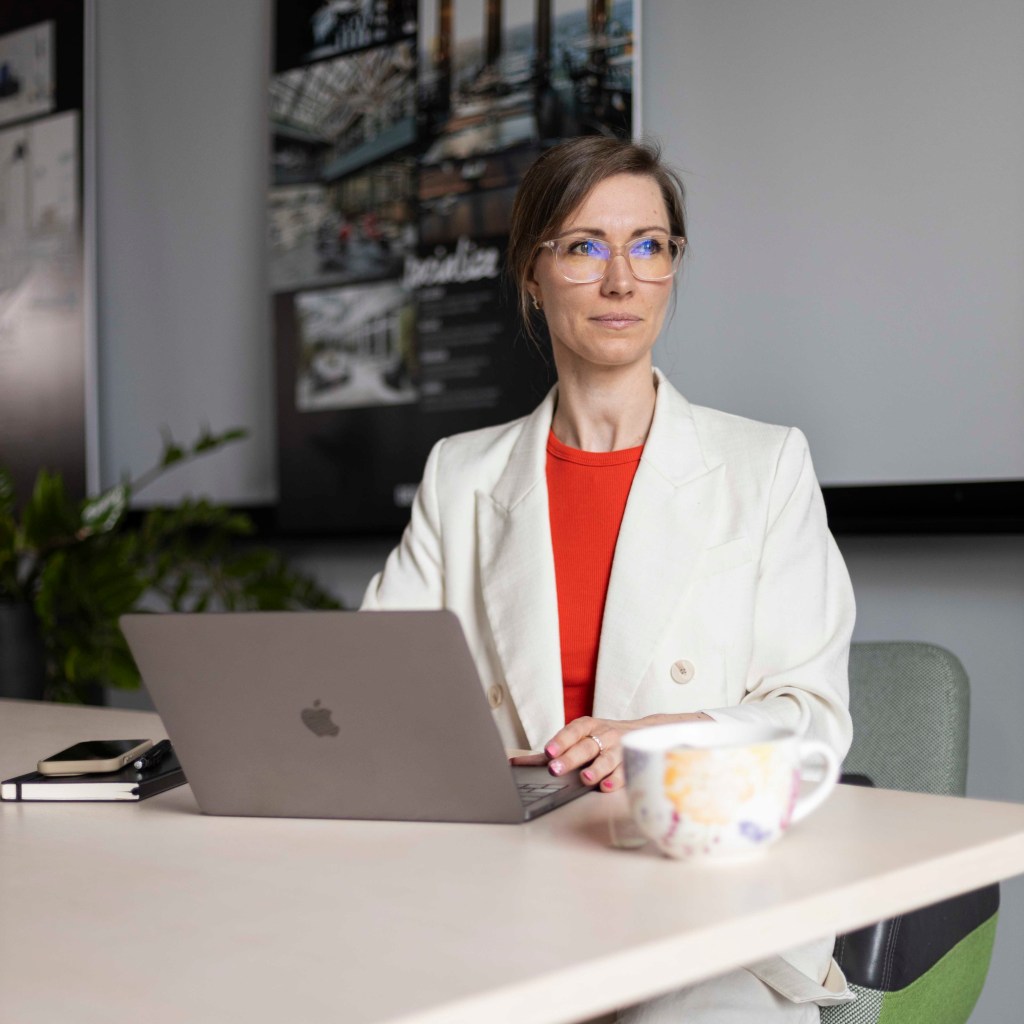 Projektmanagerin Janine Frank sitzt mit einem hellen Blazer und einem roten Shirt an einem Schreibtisch. Vor ihr Lap Top, Handy und Notizbuch. Sie schaut nach rechts.